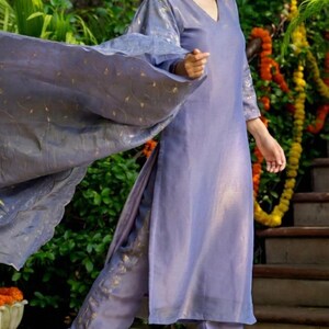 May include: A periwinkle-colored, two-piece outfit with gold accents. The ensemble includes a long-sleeved tunic and matching pants. A flowing scarf with gold detailing is draped over the shoulder. The outfit is worn with gold-colored flats.