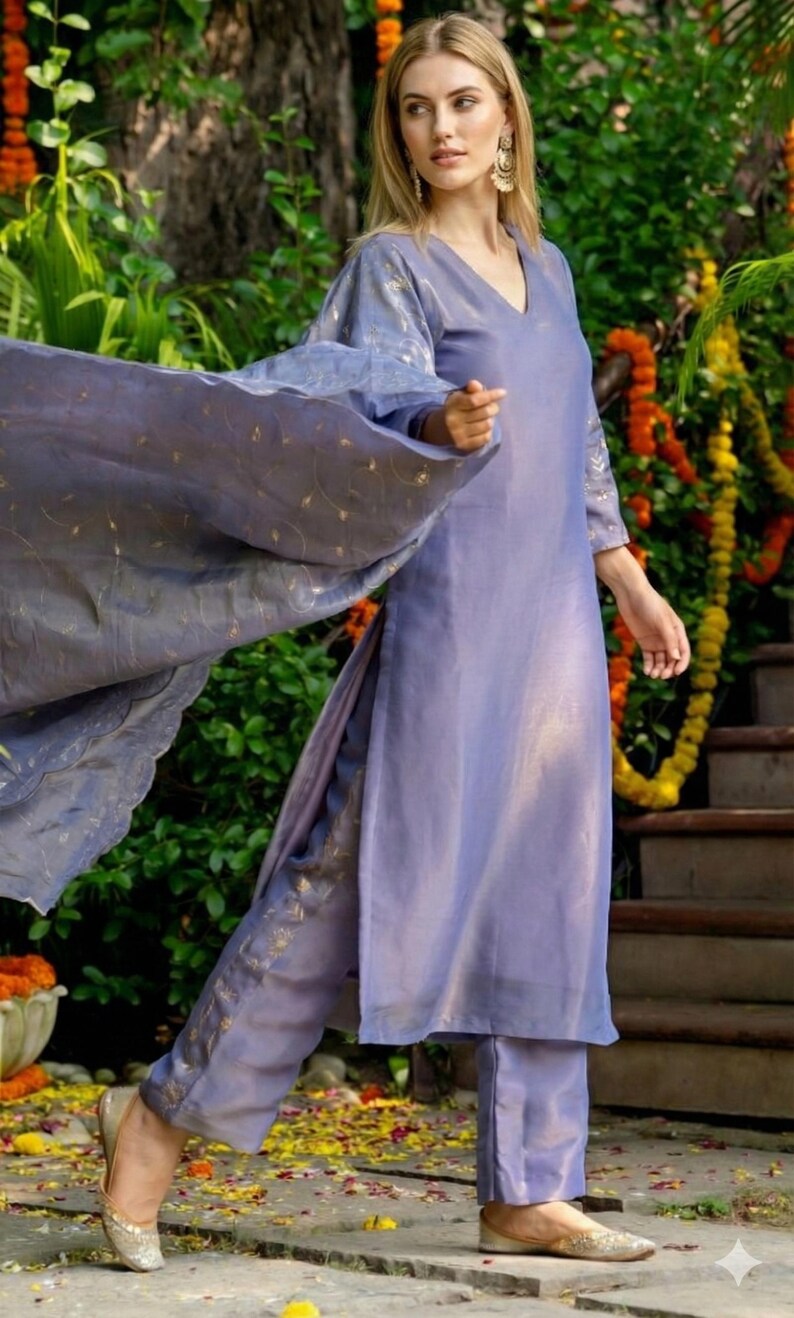 May include: A periwinkle-colored, two-piece outfit with gold accents. The ensemble includes a long-sleeved tunic and matching pants. A flowing scarf with gold detailing is draped over the shoulder. The outfit is worn with gold-colored flats.