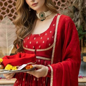 May include: A vibrant red long-sleeved dress featuring a subtle gold floral pattern, a scoop neckline, and gold trim detailing. The garment is paired with a matching red dupatta and a white beaded choker necklace with a central green stone.