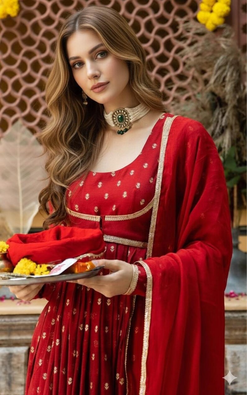 May include: A vibrant red long-sleeved dress featuring a subtle gold floral pattern, a scoop neckline, and gold trim detailing. The garment is paired with a matching red dupatta and a white beaded choker necklace with a central green stone.