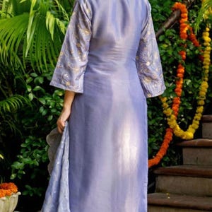 May include: A periwinkle-colored salwar kameez ensemble. The long tunic features gold embroidery on the sleeves. The matching pants and scarf complete the outfit. The model is standing in front of greenery and stairs.