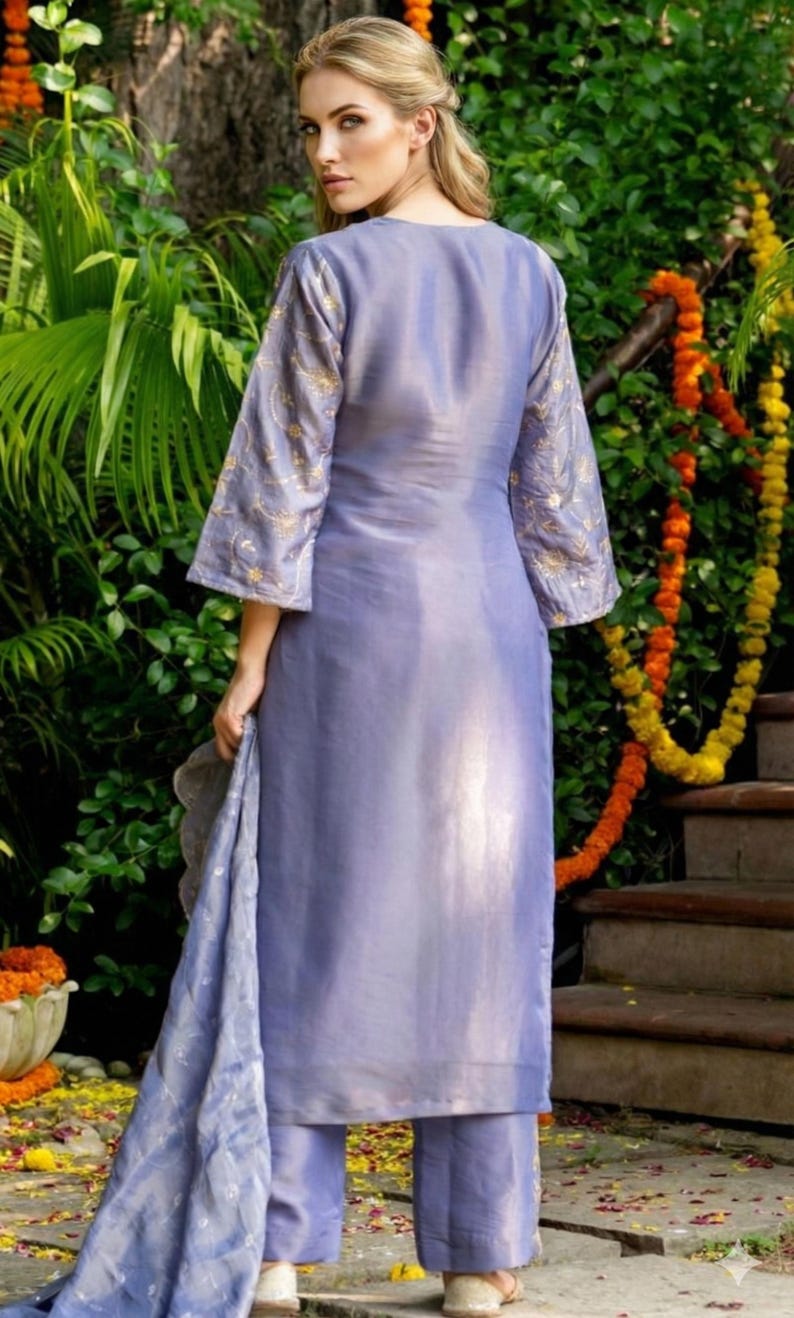 May include: A periwinkle-colored salwar kameez ensemble. The long tunic features gold embroidery on the sleeves. The matching pants and scarf complete the outfit. The model is standing in front of greenery and stairs.