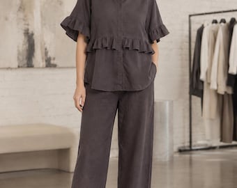 May include: A dark charcoal gray two-piece set consisting of a button-down shirt with short ruffled sleeves and a ruffled waist detail, paired with matching wide-leg trousers featuring three small white buttons at the bottom of the right leg.