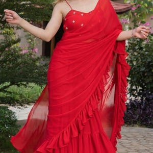 May include: A flowing red saree with a ruffled hem and a matching top with delicate strap details. The saree is draped elegantly, creating a dynamic silhouette. The fabric appears lightweight and airy, perfect for special occasions.