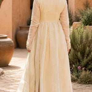 May include: A cream-colored long-sleeved tunic with a fitted bodice and a pleated, flowing skirt, paired with matching wide-leg trousers. The garment features sheer fabric overlays and subtle metallic embroidery details on the sleeve cuffs.