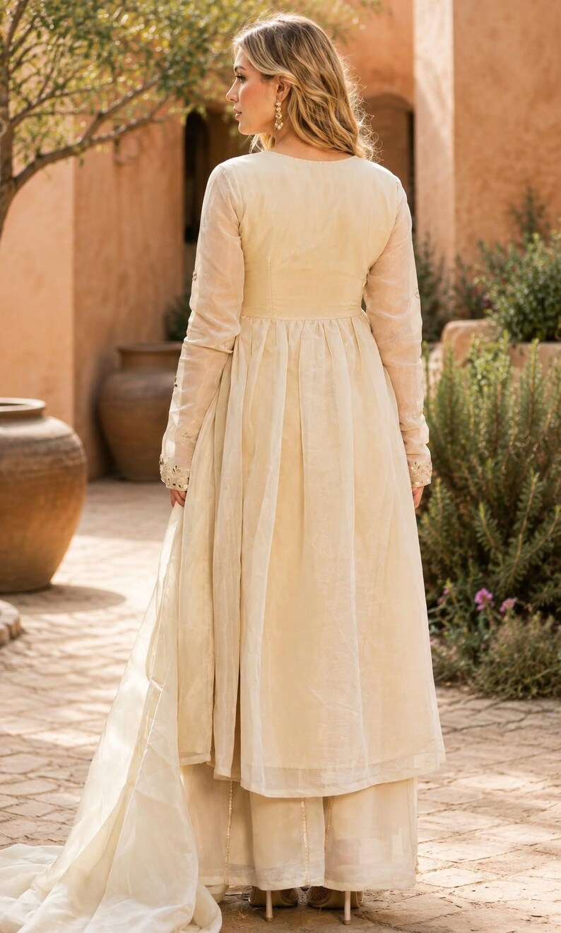 May include: A cream-colored long-sleeved tunic with a fitted bodice and a pleated, flowing skirt, paired with matching wide-leg trousers. The garment features sheer fabric overlays and subtle metallic embroidery details on the sleeve cuffs.