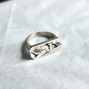 May include: A silver ring with a rectangular band featuring an engraved design of birds in flight.
