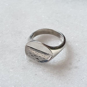 May include: A silver signet ring with an oval face and a textured design. The ring is displayed on a white marble surface, showcasing its polished metal finish. This piece of jewellery is suitable for everyday wear.