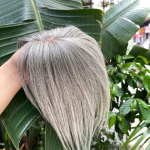 以下が含まれることがあります： 自然な頭皮を模したベースに、ストレートのシルバーグレーの髪をあしらったヘアトッパー。髪は滑らかでレイヤーが入っており、明るいグレーと暗いグレーが混ざり合っています。頭頂部をカバーするためにデザインされたヘアピースです。