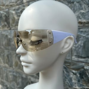 May include: Stylish, rimless sunglasses with a light purple frame and tinted lenses. The sunglasses have a sleek, modern design with silver-colored rivets on the temples. The lenses are a light brown color.