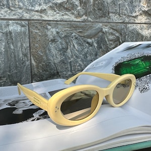 Puede incluir: Gafas de sol ovaladas color crema con lentes oscuras sobre un libro abierto. Las gafas tienen una montura gruesa y un detalle rectangular en las patillas. El fondo presenta una pared de piedra y una revista.