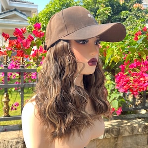 Op de afbeelding: Een bruine baseballcap met een gebogen klep, gedragen op een mannequinhoofd met golvend bruin en blond haar. De cap heeft een klein wit geborduurd ontwerp op de voorkant. De achtergrond toont levendige roze bloemen en groen.