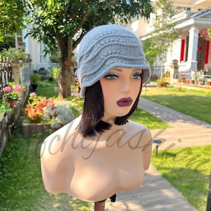 May include: A light gray knitted cloche hat with a slightly curved brim. The hat has a textured, ribbed design and is displayed on a mannequin head. The background features a lush green yard and a house.