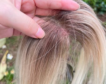 Könnte beinhalten: Ein Haarteil mit einer Monofilament-Basis und einem sichtbaren Gittermuster. Das Haar verläuft von einem dunkelbraunen Ansatz an der Basis zu einem helleren, aschblonden Farbton in den Längen und zeigt einen natürlich wirkenden Farbverlauf.