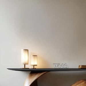 May include: Two cylindrical table lamps with beige shades and brass bases are illuminated on a black table. The word "Width" is displayed in silver letters. The table has a dark wood base.