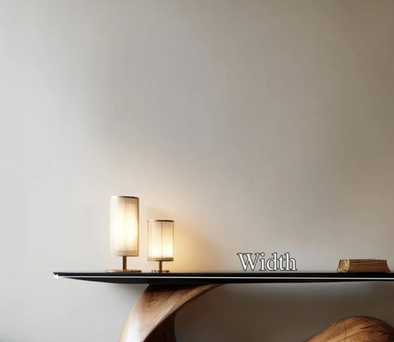 May include: Two cylindrical table lamps with beige shades and brass bases are illuminated on a black table. The word "Width" is displayed in silver letters. The table has a dark wood base.