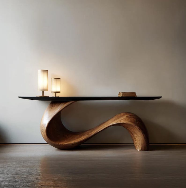 May include: A contemporary wooden console table with a distinctive, curved design. The table has a dark, rectangular top and a sculptural base crafted from light brown wood. Two small lamps with cylindrical shades are on the table, and a small book rests on the right side.