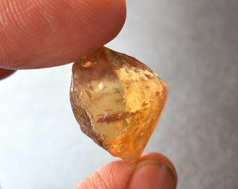 Op de afbeelding: Een ruwe, amberkleurige edelsteen met meerdere facetten wordt tussen twee vingers gehouden. De edelsteen heeft een kristalstructuur en een warme, gouden tint. De steen lijkt doorschijnend, met licht dat door de facetten gaat. De achtergrond is een vage, neutrale grijstint.
