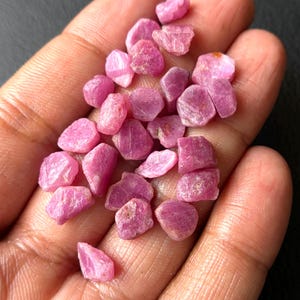 以下が含まれることがあります： 未加工のピンク色のルビーの宝石の束。未研磨の石はサイズと形状が異なり、さまざまなピンクの色合いを示しています。宝石は、暗い背景に対して開いた手で保持されています。