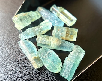 May include: A collection of raw aquamarine crystals in varying shapes and sizes. The crystals display a range of pale blue and greenish-blue hues, with a translucent quality. They are arranged on a dark, textured surface.