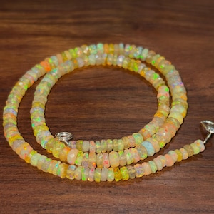 May include: A necklace composed of small, faceted, circular opal beads. The beads exhibit a translucent, milky base color with vibrant flashes of orange, green, and yellow iridescence. The strand is secured with a small, silver-toned metal clasp.