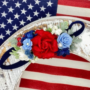 Puede incluir: Una diadema de flores roja, blanca y azul con una banda elástica azul marino. La diadema está decorada con rosas rojas, flores azules y flores blancas.