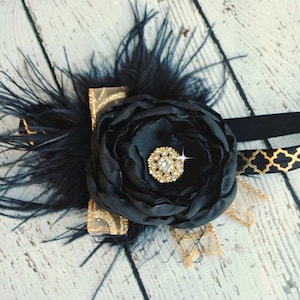 May include: Black headband with a large black satin flower, gold trim, and a black feather accent. The flower has a rhinestone center.