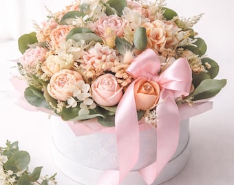 Könnte beinhalten: Eine runde, weiße Geschenkbox mit einem Blumenarrangement. Die Blumen sind in Pfirsich- und Rosatönen gehalten, mit grünem Laub und einem rosa Satinband. Die Box hat ein zartes, geprägtes Muster.