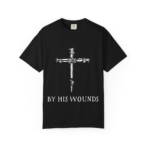 Op de afbeelding: Zwart T-shirt met een witte afbeelding van een kruis gemaakt van spijkers. De tekst "BY HIS WOUNDS" is onder het kruis gedrukt in een schreefletter. Het shirt is gemaakt van een zacht materiaal en heeft korte mouwen.