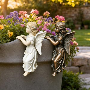 May include: Two angel figurines perched on the rim of a large gray planter. One angel is finished in a matte white, while the other has a metallic bronze finish. Both figures feature detailed wings, flowing robes, and hair adorned with a small headband.