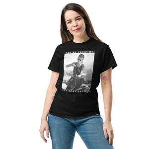 Może przedstawiać: Czarny t-shirt z czarno-białym zdjęciem osoby i napisem "ASK ME ABOUT MY FEMINIST AGENDA". Osoba ma na sobie okulary przeciwsłoneczne i trzyma broń.