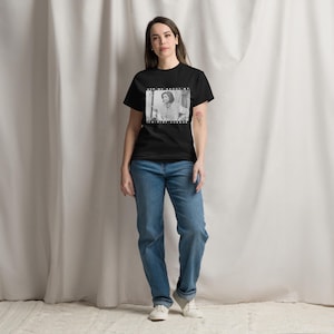 Puede incluir: Una camiseta negra con una imagen en blanco y negro de una mujer y el texto "ASK ME ABOUT MY FEMINIST AGENDA". La persona lleva vaqueros azules y zapatillas blancas.