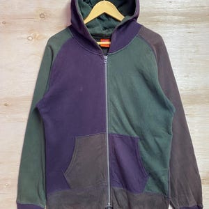 Peut inclure: Un sweat à capuche zippé multicolore avec une capuche. Le sweat à capuche présente un design color-block avec des nuances de vert, de violet et de marron. Il possède une poche avant et une fermeture éclair intégrale. Le sweat à capuche est suspendu à un cintre en bois.