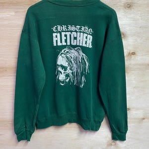May include: A dark green sweatshirt with the name "CHRISTIAN FLETCHER" printed in white block letters above a white skull graphic with long hair. The sweatshirt is hanging on a wooden hanger.