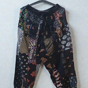 May include: Black patchwork trousers with a drawstring waist, hanging on a black hanger. The trousers feature a variety of patterned fabric squares in colours like red, gold, and white. The overall style is bohemian.