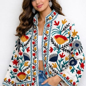 Puede incluir: Chaqueta bordada color crema con cuello alto. La chaqueta presenta un vibrante diseño floral en rojo, amarillo, azul y verde. El bordado está delineado con un borde turquesa. La chaqueta se combina con unos vaqueros azules.