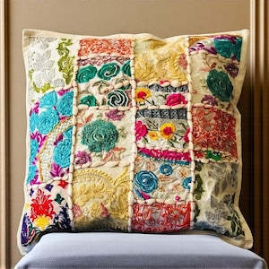 Puede incluir: Cojín decorativo cuadrado con diseño de patchwork. Presenta patrones florales bordados en colores como turquesa, rosa, amarillo y rojo sobre un fondo crema. El cojín está colocado sobre una superficie azul claro. Es un artículo decorativo.