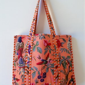 Puede incluir: Bolso tote acolchado con un vibrante estampado floral y de pájaros en tonos coral, rojo, azul y verde. El bolso tiene asas largas y está ribeteado con tela a rayas naranja y blanca. Ideal para llevar objetos.