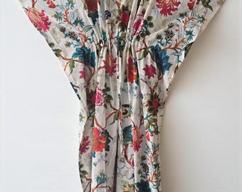 May include: A white caftan with a vibrant floral pattern. The design features large, colorful flowers in shades of red, pink, blue, and green, set against a neutral background. The caftan has a V-neck and a loose, flowing silhouette.