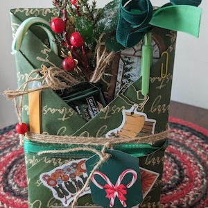 May include: A green gift-wrapped package featuring gold cursive text, adorned with a candy cane, red berries, a green velvet ribbon, a light green pen, a gold paperclip, and stickers depicting books, flowers, and two crossed candy canes.