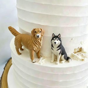 May include: A multi-tiered white cake with horizontal ridges, topped with two dog figurines. One figurine is a standing golden-brown dog, and the other is a sitting black, white, and gray husky-type dog, both placed on the white frosting surface.