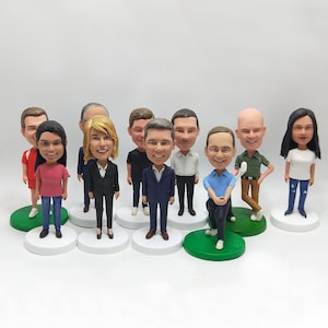 Può includere: Una collezione di nove statuette bobblehead personalizzate posizionate su basi circolari individuali. Ogni statuetta raffigura una persona unica con tratti del viso, acconciature e abbigliamento distinti, tra cui camicie casual, completi e un golfista con mazza.