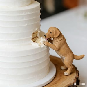 May include: A three-tiered white cake with textured frosting, with a bite taken out of the side. A tan-colored dog figurine is reaching up to eat the cake, with its tongue sticking out. The cake sits on a wooden base.