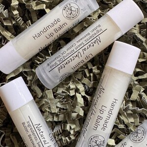 May include: Four cylindrical tubes of handmade lip balm with white caps and clear plastic bodies. Each label features the text Handmade Lip Balm, Natural Unscented, and a circular logo with a goat illustration. Ingredients include sweet almond oil.