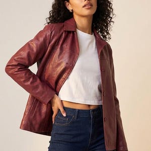 Puede incluir: Chaqueta de cuero burdeos con cuello y cierre de botones. La chaqueta se combina con una camiseta blanca y unos vaqueros azul oscuro. La persona lleva pendientes pequeños dorados.