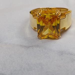 May include: A gold-toned ring featuring a large, rectangular, faceted yellow gemstone set in a four-prong setting. The band is wide and smooth, with a polished metallic finish that complements the vibrant, transparent yellow stone at the center.