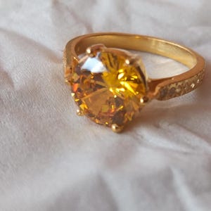 May include: A gold ring featuring a large, faceted, yellow-orange gemstone. The ring's band has a textured pattern on the sides. The gemstone is central, reflecting light and displaying shades of yellow and orange. A statement piece.