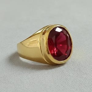 May include: A gold ring featuring a large, oval-cut, deep red gemstone. The ring has a thick, polished gold band and a bezel setting that securely holds the gemstone. The gemstone's facets catch the light, creating a brilliant display.
