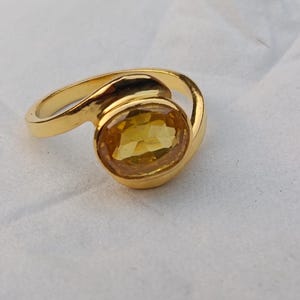 May include: A gold ring featuring a large, oval yellow gemstone. The ring's band curves around the gemstone, creating an elegant design. The gemstone is a bright, warm yellow colour, and the ring is set against a white background.