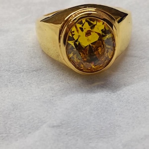May include: A gold ring featuring an oval yellow gemstone. The ring has a substantial band and a bezel setting. The gemstone is multi-faceted, creating a sparkling effect. The ring is likely made of gold-plated metal.